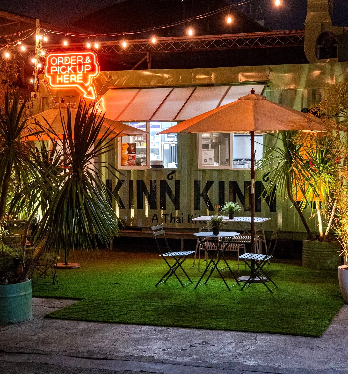 The Kinn Kinn green container kitchen at dusk, with umbrellas, string lights and a neon order sign